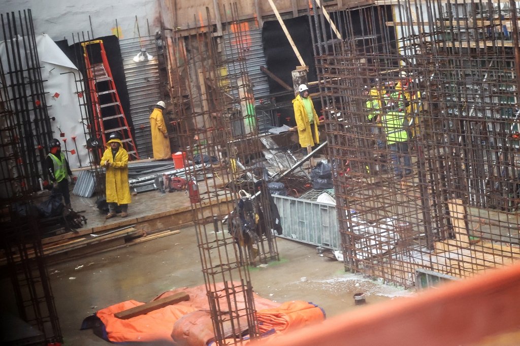 A construction site with workers m moving around in the rain with lots of rebar around