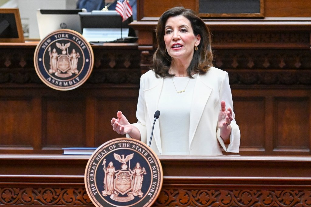 Hochul in white giving her 2023 State of the State address from within the wood-paneled Assembly chambers in Albany